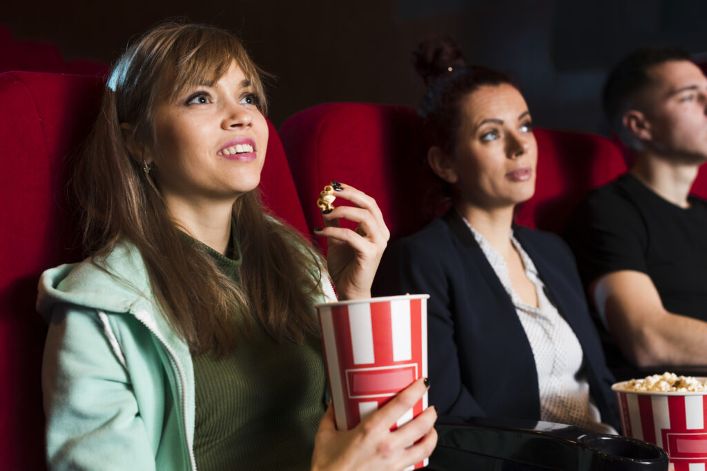 Three young people enjoying a movie night at the best cinema, sitting in red theater seats with popcorn. A smiling woman in a green hoodie holds a striped popcorn bucket and looks up at the screen, while her friends watch attentively beside her.