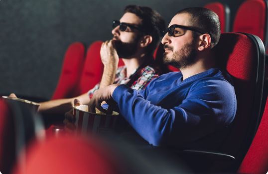 Two men enjoying a movie at the best cinema, sitting in red theater seats while wearing 3D glasses. The man in a blue sweater holds a popcorn bucket and looks focused on the screen, while his friend in a plaid shirt eats popcorn beside him.
